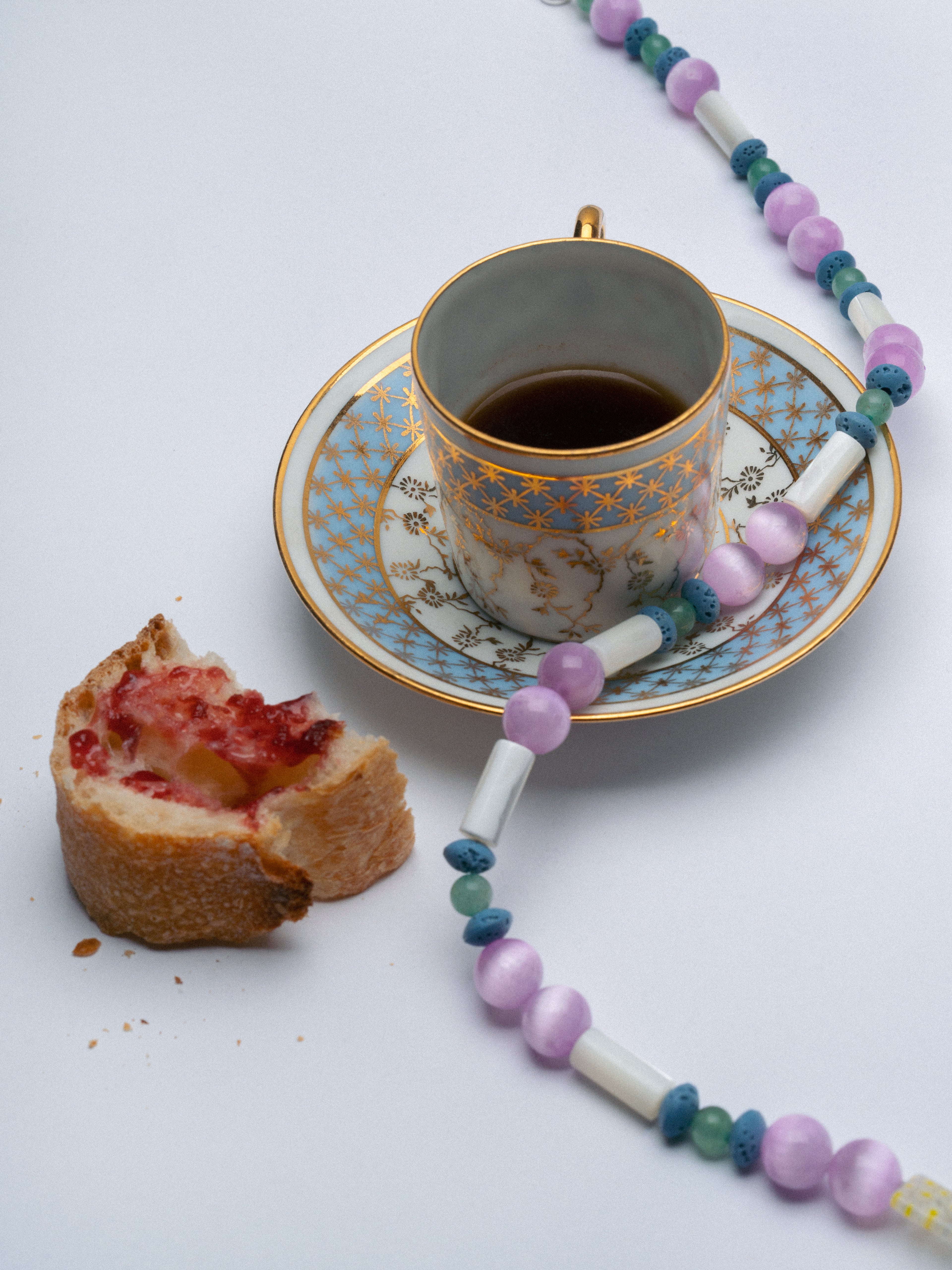 A styled flat-lay featuring a delicate beaded necklace with lavender, teal, and white beads draped around a gold-patterned teacup and saucer, with a half-eaten pastry on a white background.
